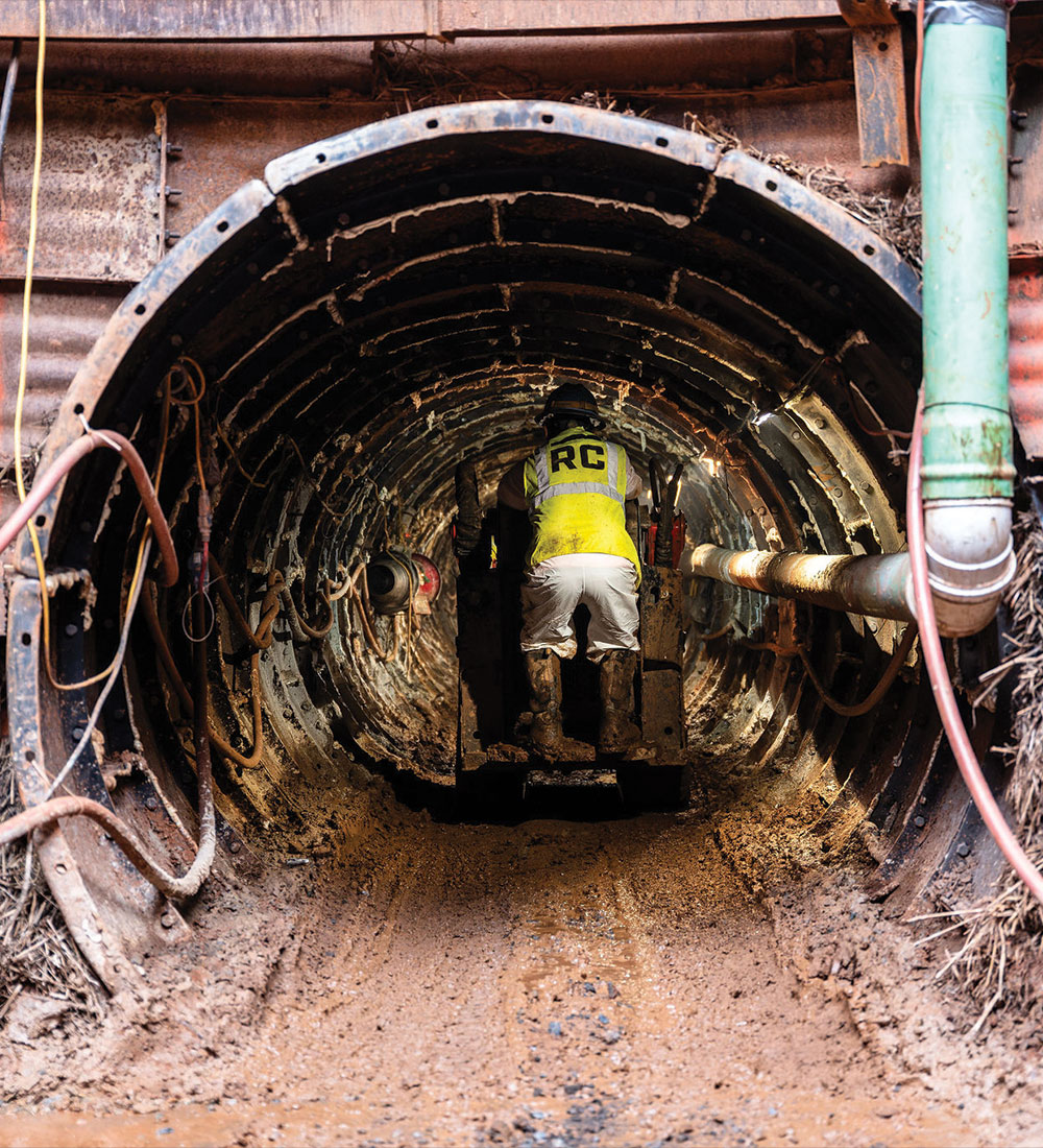 Worker in RC vest operates machinery in muddy, unfinished tunnel.