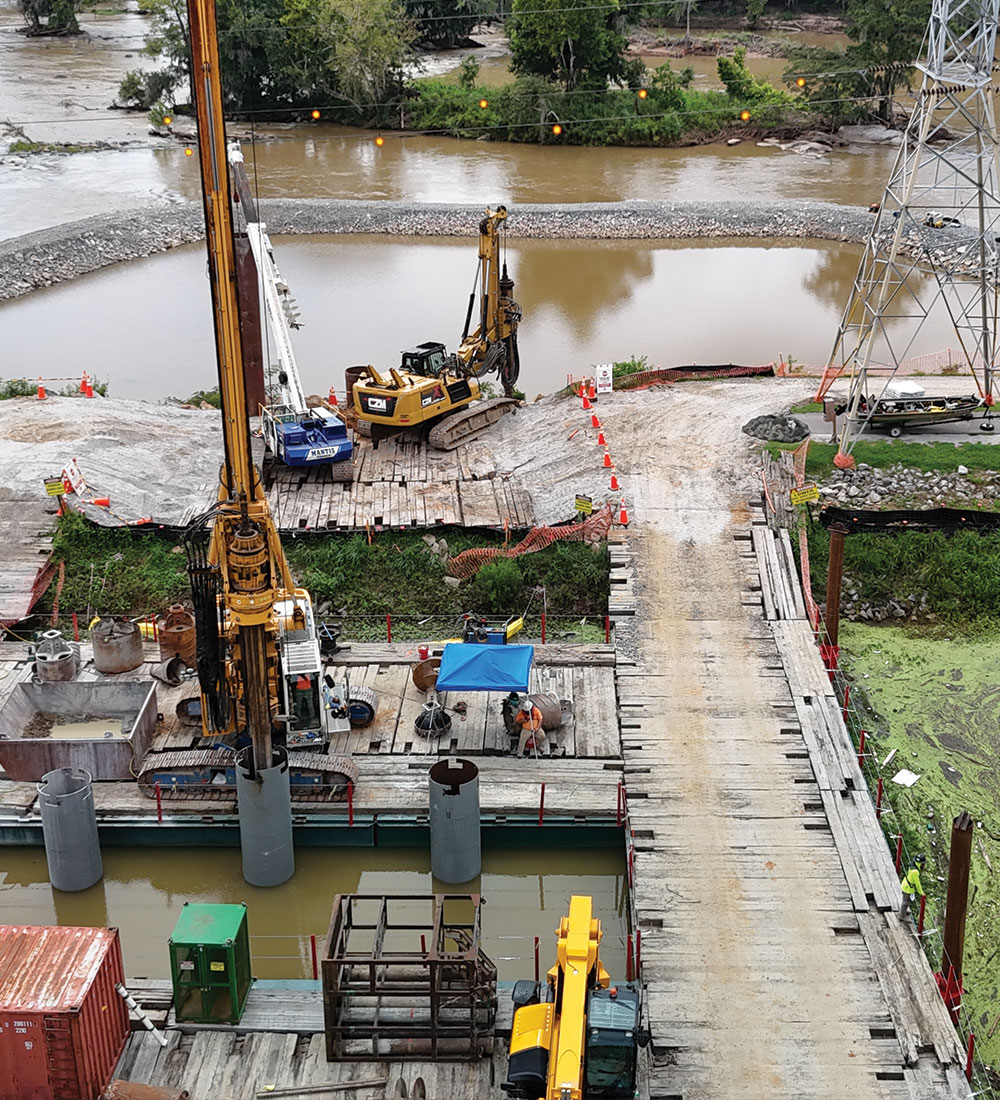 Obra junto a la orilla del río con maquinaria, trabajadores, taladros y una pasarela de madera.