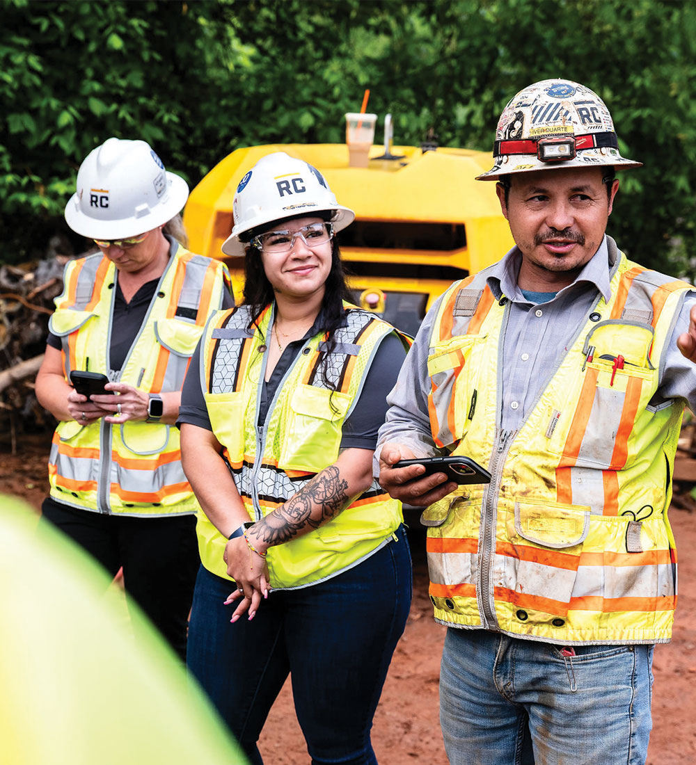 Three construction workers stand outside; two use phones, one gestures.
