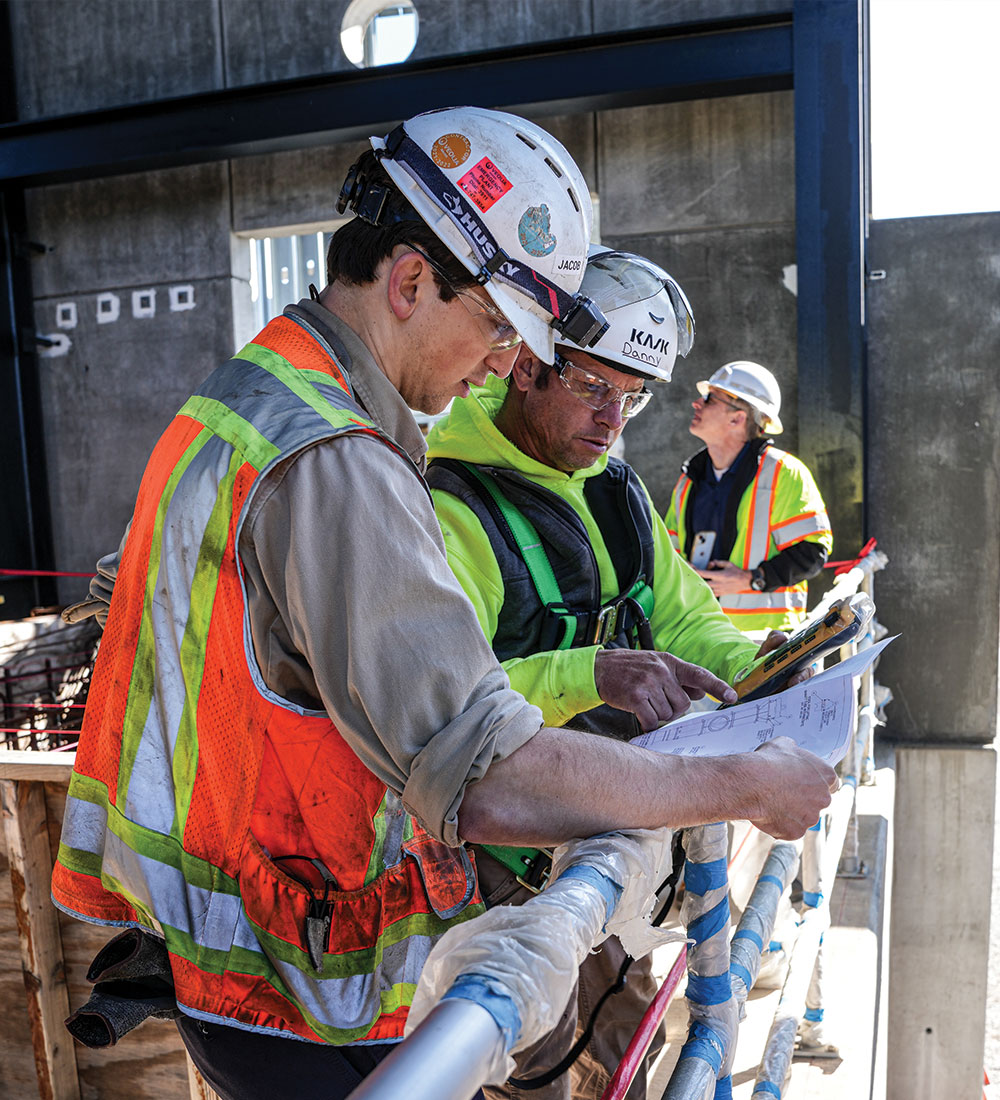 Construction workers in safety gear review blueprints; one observes.
