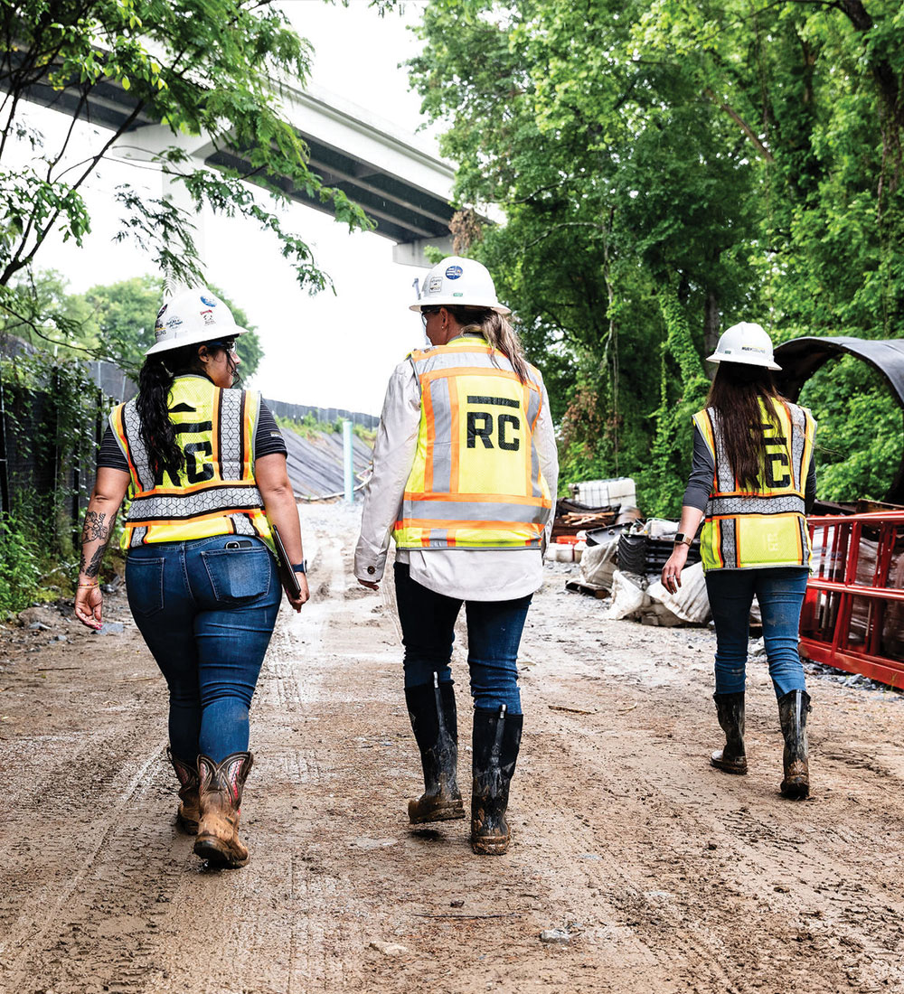 Three workers in safety vests walk on muddy site path.