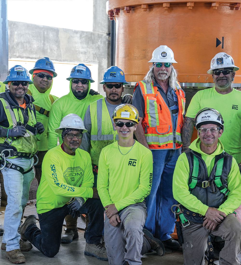 Construction workers in safety gear pose by industrial equipment.