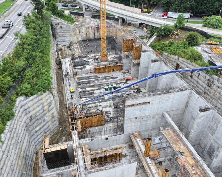 Aerial view: construction site, crane, workers, highway, green trees.