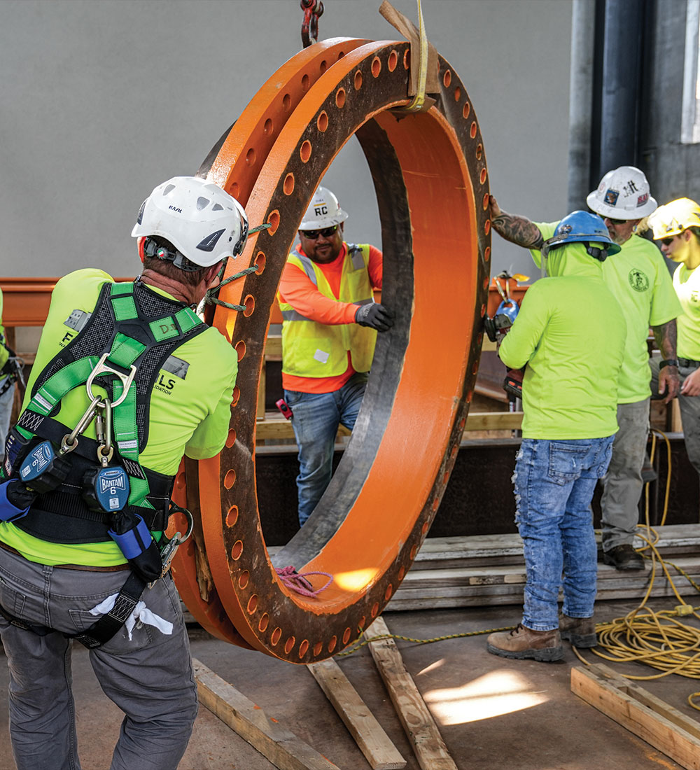 Workers in safety gear position a large orange metal ring indoors.