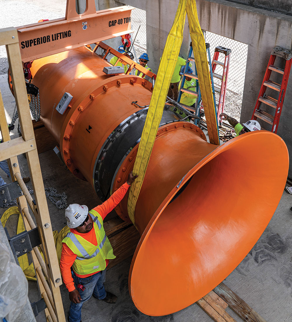 Workers lift large orange pipe with crane at construction site.
