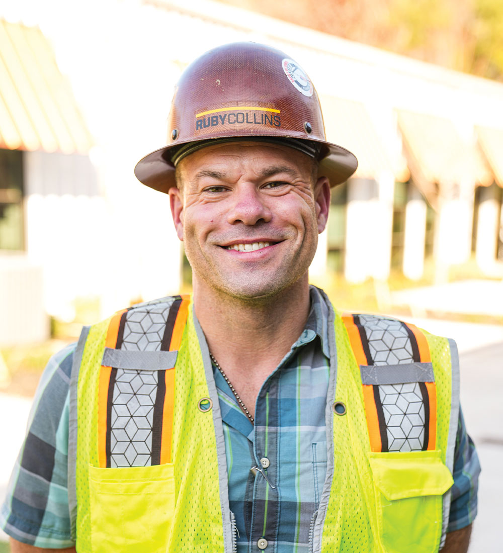Smiling worker in RUBY COLLINS hard hat and vest outside.