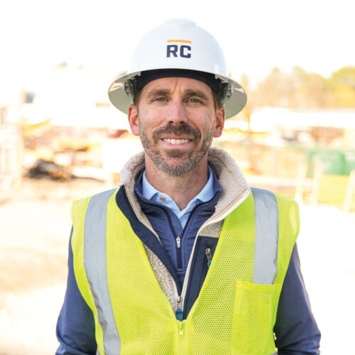 Smiling man in RC hard hat and yellow vest at construction site.