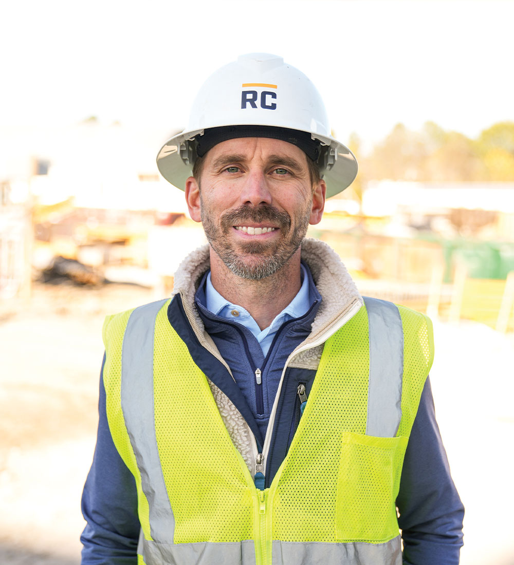 Smiling man in RC hard hat and yellow vest at construction site.