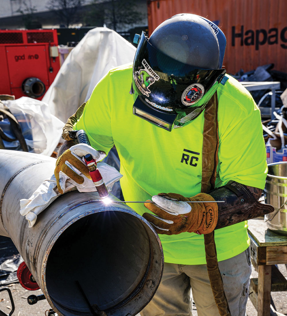 Worker in yellow shirt welding a metal pipe outdoors.