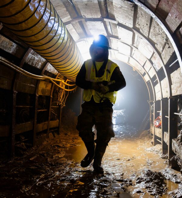 Worker in safety gear stands in a muddy, dimly lit tunnel.