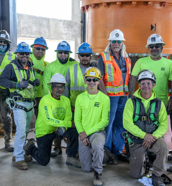Workers in safety gear pose by a large orange industrial part.