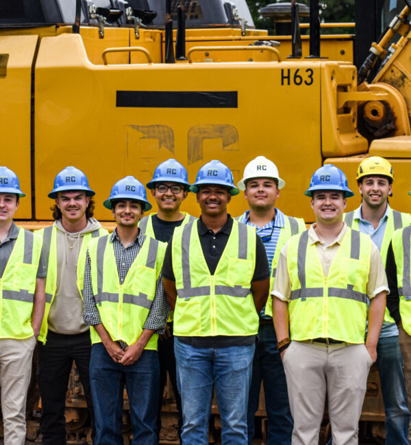 Eleven workers in vests and hard hats by yellow vehicle.