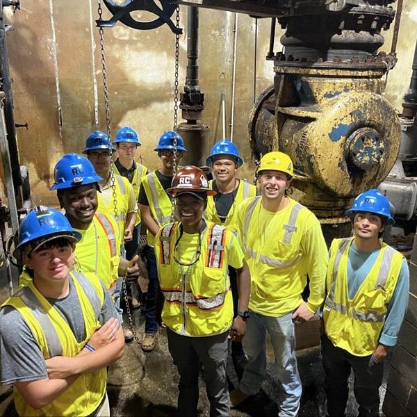 Construction workers in helmets and vests stand by industrial pipes.