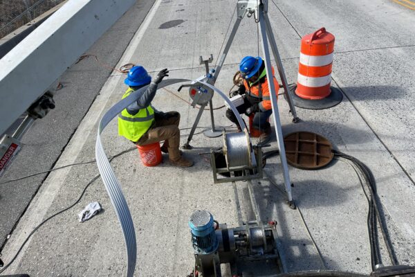 Unos trabajadores, equipados con material de seguridad, manejan la maquinaria y guían el cable en el puente.