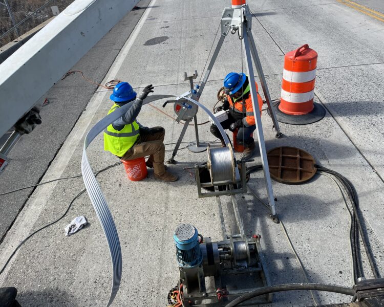 Workers in safety gear operate equipment and guide cable on bridge.