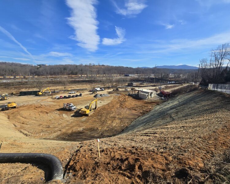 Excavators and vehicles at a dirt construction site, mountains behind.
