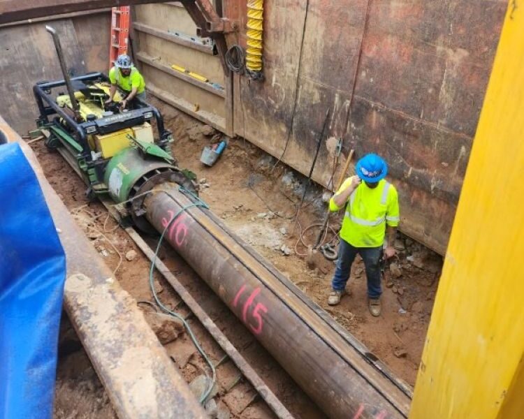 Two workers install a steel pipe using machinery at a dig site.