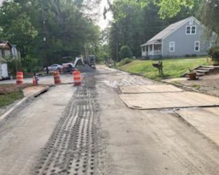 Calle en obras con tierra, huellas de neumáticos, tablones y barriles.