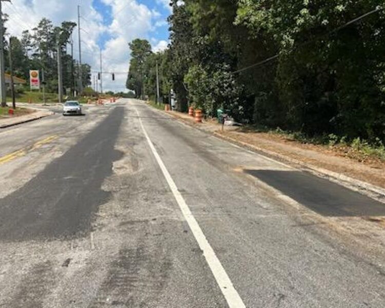 Carretera parcialmente repavimentada, conos, coche blanco a lo lejos, árboles a la derecha.
