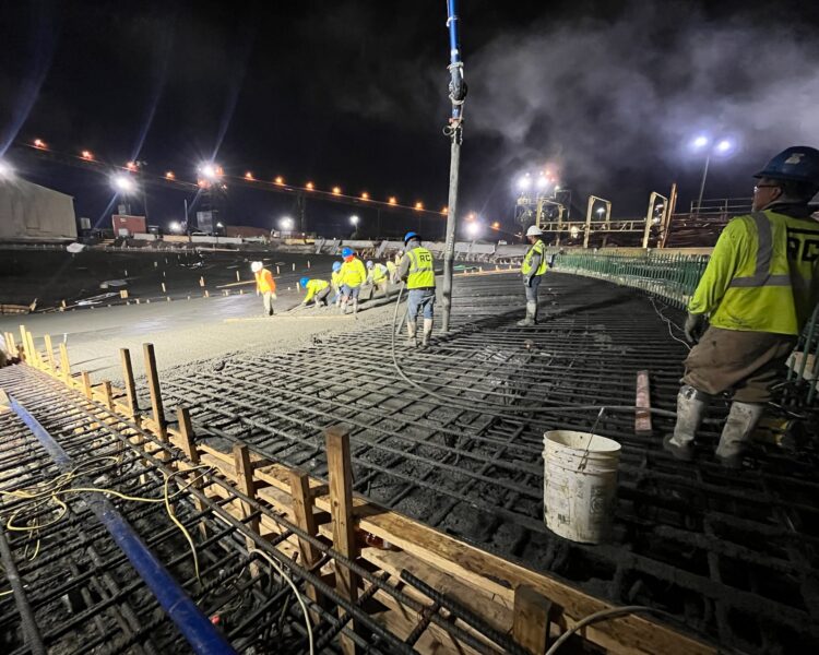 Workers pour concrete at night under bright lights; rebar visible.