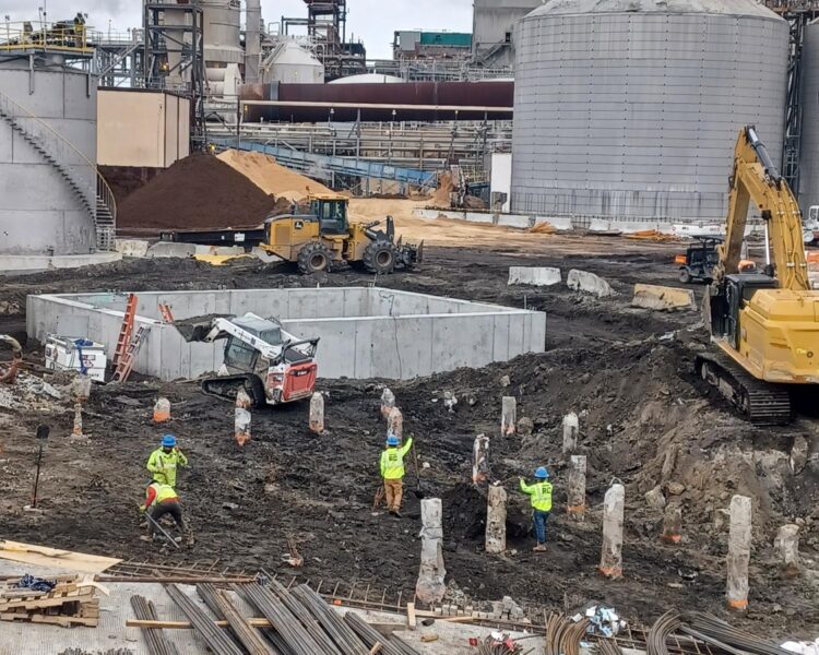 Workers and machines on a muddy site with storage tanks.