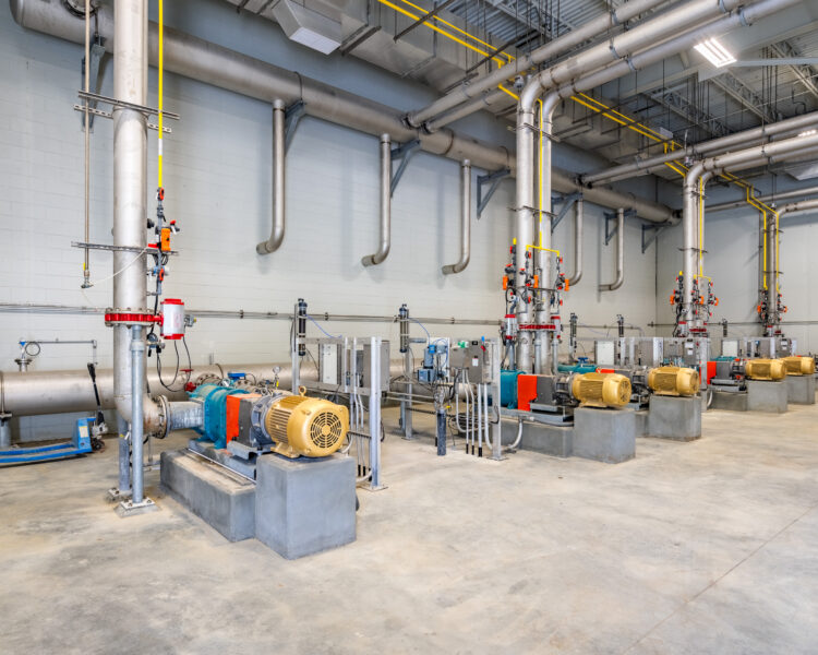 Nave industrial con bombas, tuberías sobre plataformas y cableado a la vista.