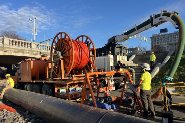 Unos trabajadores utilizan maquinaria y carretes de cable de color naranja junto a un puente.