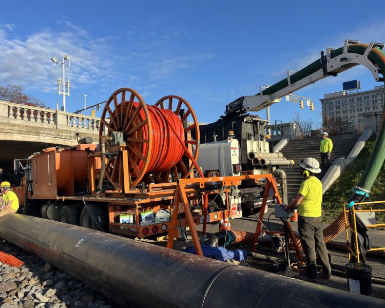 Unos trabajadores utilizan maquinaria y carretes de cable de color naranja junto a un puente.
