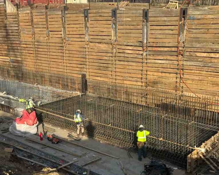 Workers install rebar in a foundation pit by shipping containers.