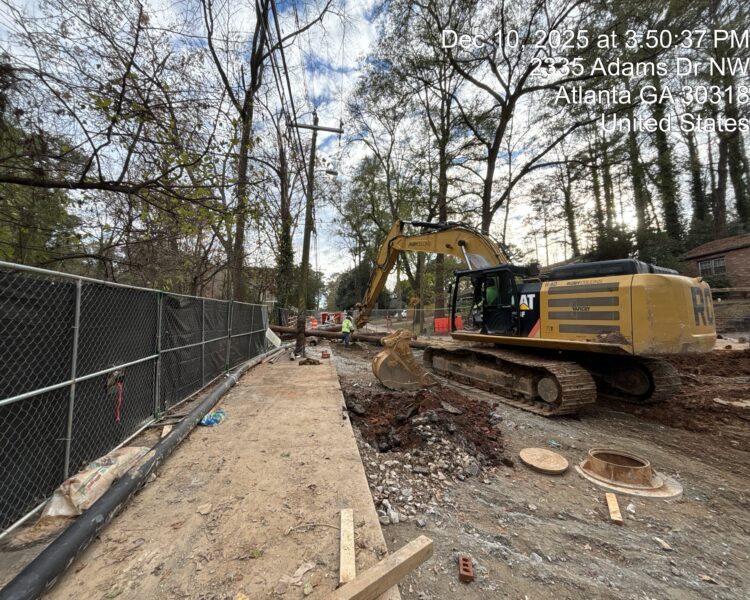 Excavator at construction site near fence; workers, trees, text overlay.
