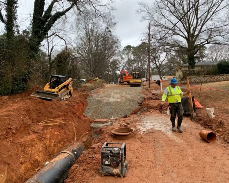 Los trabajadores y la maquinaria colocan tuberías y grava en una carretera embarrada.