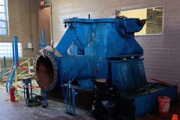 Bomba industrial azul oxidada entre baldosas, cinta de señalización y una bolsa de basura.