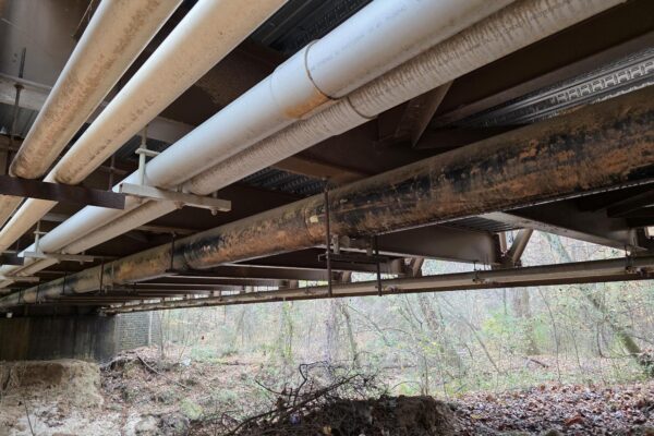 Las tuberías discurren en paralelo bajo un puente que se eleva sobre un terreno boscoso y cubierto de escombros.