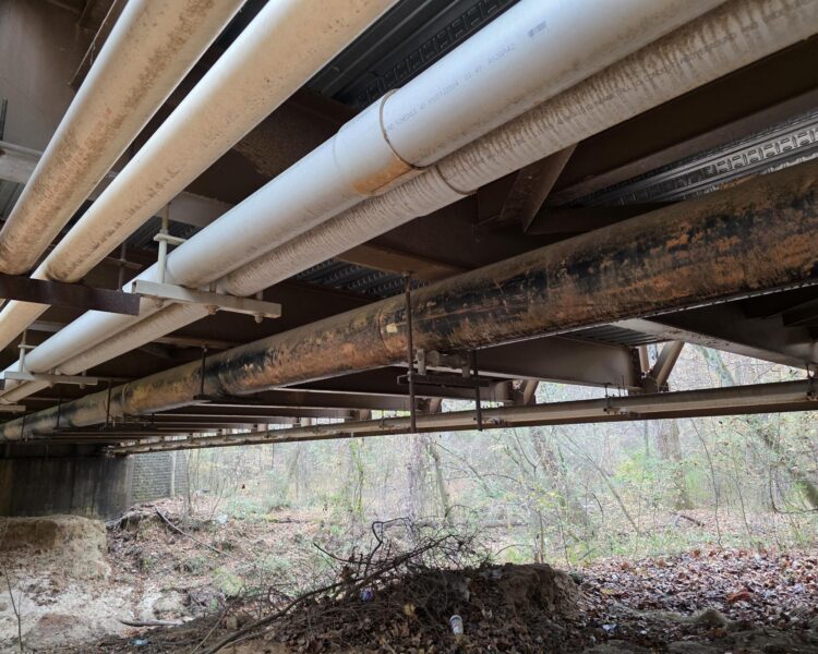 Las tuberías discurren en paralelo bajo un puente que se eleva sobre un terreno boscoso y cubierto de escombros.