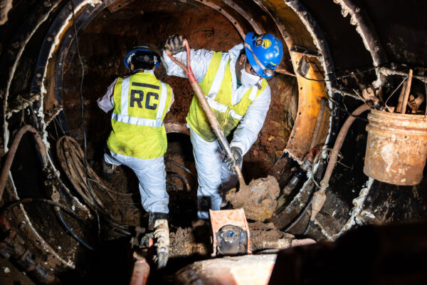 Two workers in safety gear shovel debris inside a tunnel.