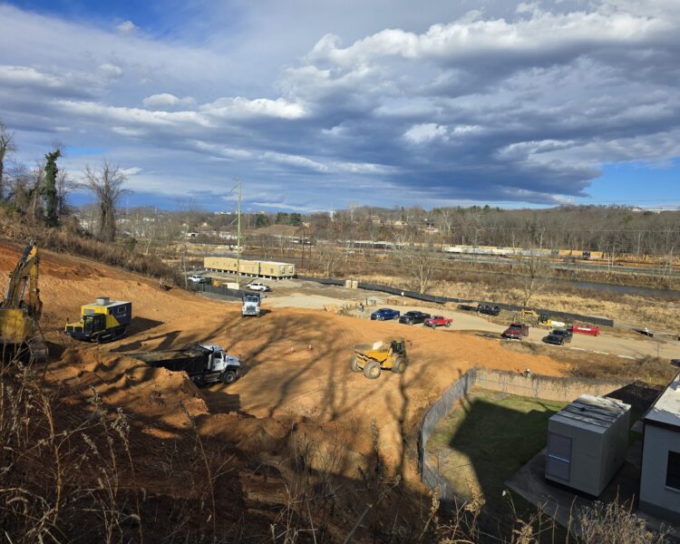 Excavators and trucks at construction site with buildings in background.