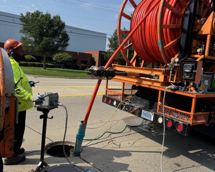 Worker feeds red hose from truck into street manhole.