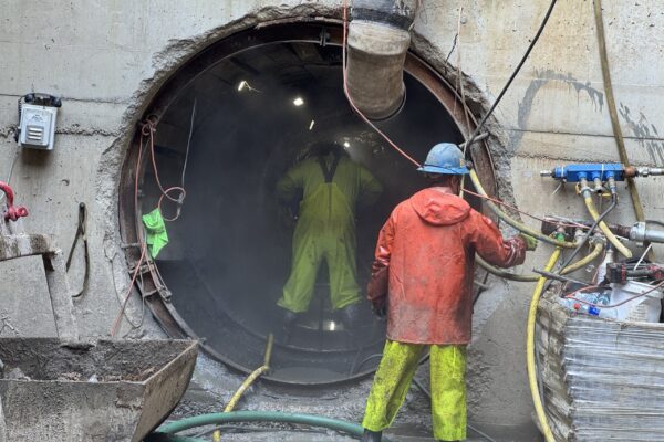Dos obreros, ataviados con su equipo de trabajo, se encuentran junto a un gran túnel de hormigón.