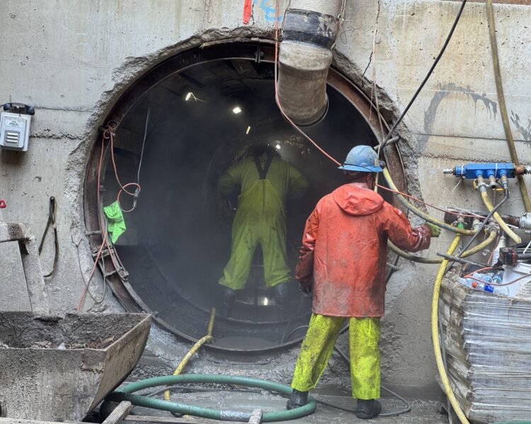 Dos obreros, ataviados con su equipo de trabajo, se encuentran junto a un gran túnel de hormigón.