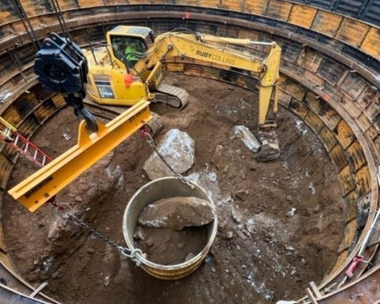 Una excavadora amarilla introduce un tubo de hormigón en un pozo circular armado.
