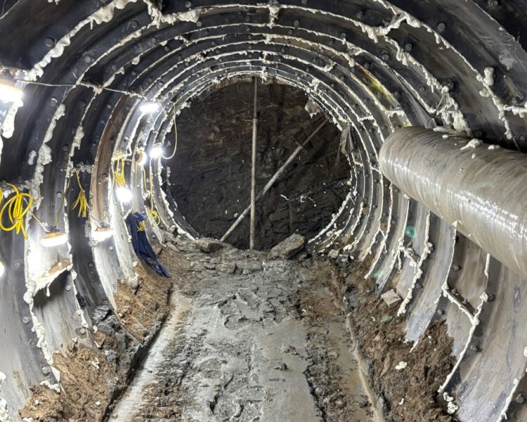 Túnel en construcción con pilares, tuberías, luces y suelo embarrado.
