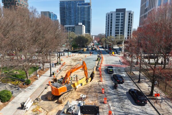 City street under construction with excavator, barrels, cars, buildings.