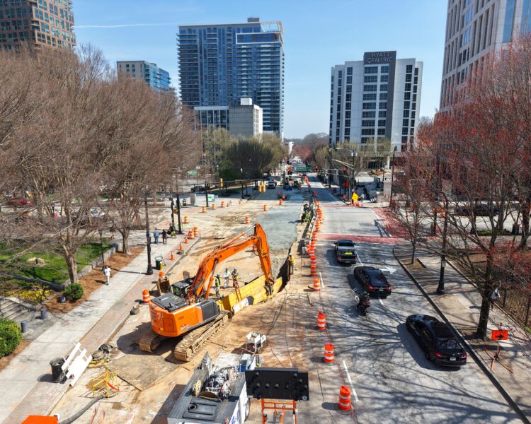 Calle de la ciudad en obras, con una excavadora, barriles, coches y edificios.