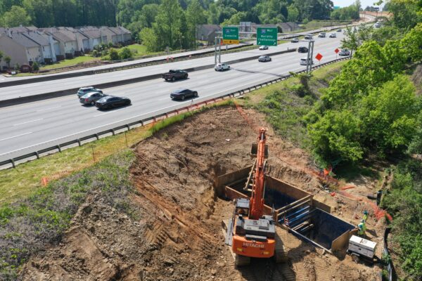 Una excavadora excava junto a la autopista; dos trabajadores manipulan materiales cerca de allí.
