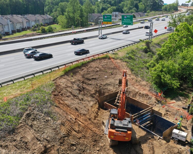 Una excavadora excava junto a la autopista; dos trabajadores manipulan materiales cerca de allí.