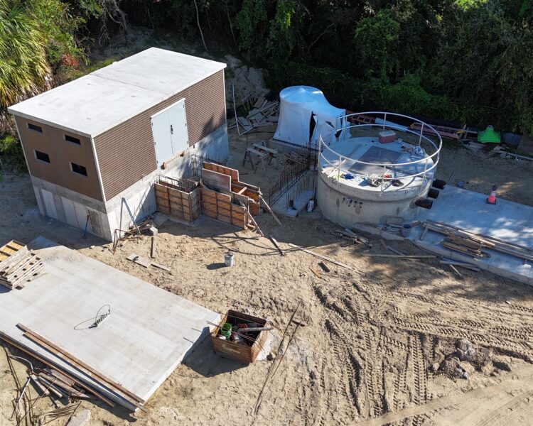 Aerial view: rectangular building, circular structure, materials on sand.