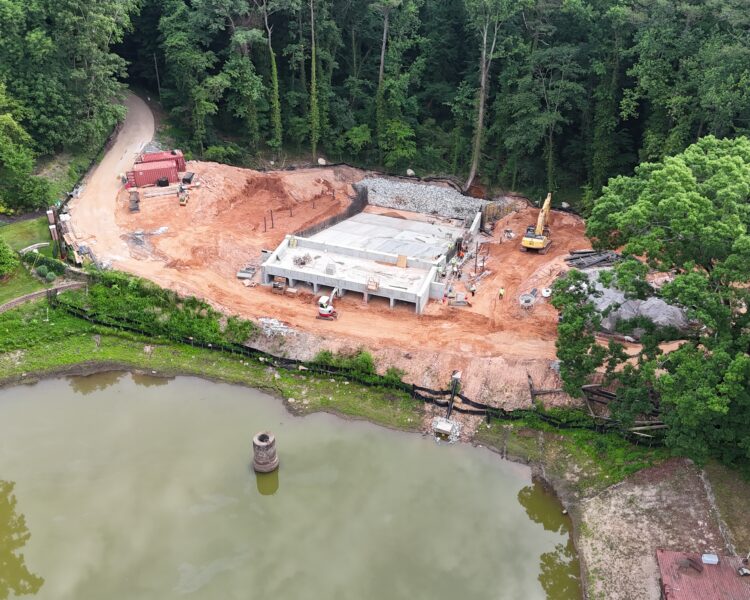 Obra junto a un estanque y un bosque, con vehículos y tierra roja.