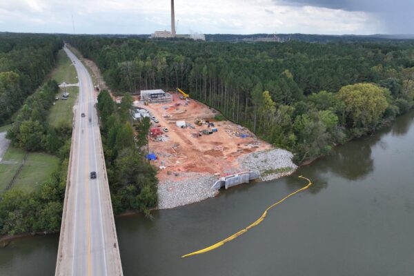 Vista aérea: obra junto al río, carretera, árboles y una instalación en la lejanía.