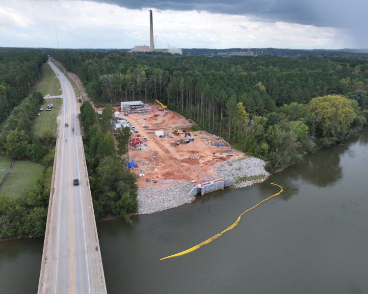 Aerial view: construction site by river, road, trees, distant facility.