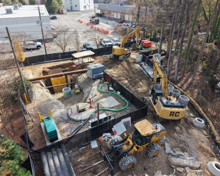 Construction site with machinery, pipes, fences; trees and buildings nearby.